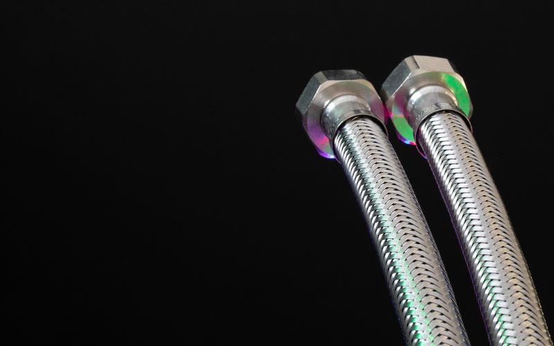 Deux flexibles à tresse en acier inoxydable et leurs écrous de raccordement, sur fond noir