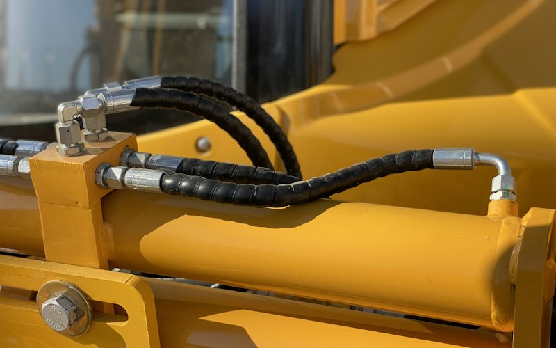 Flexibles hydrauliques noirs à raccords métalliques coudés, montés sur un vérin jaune d'engin
